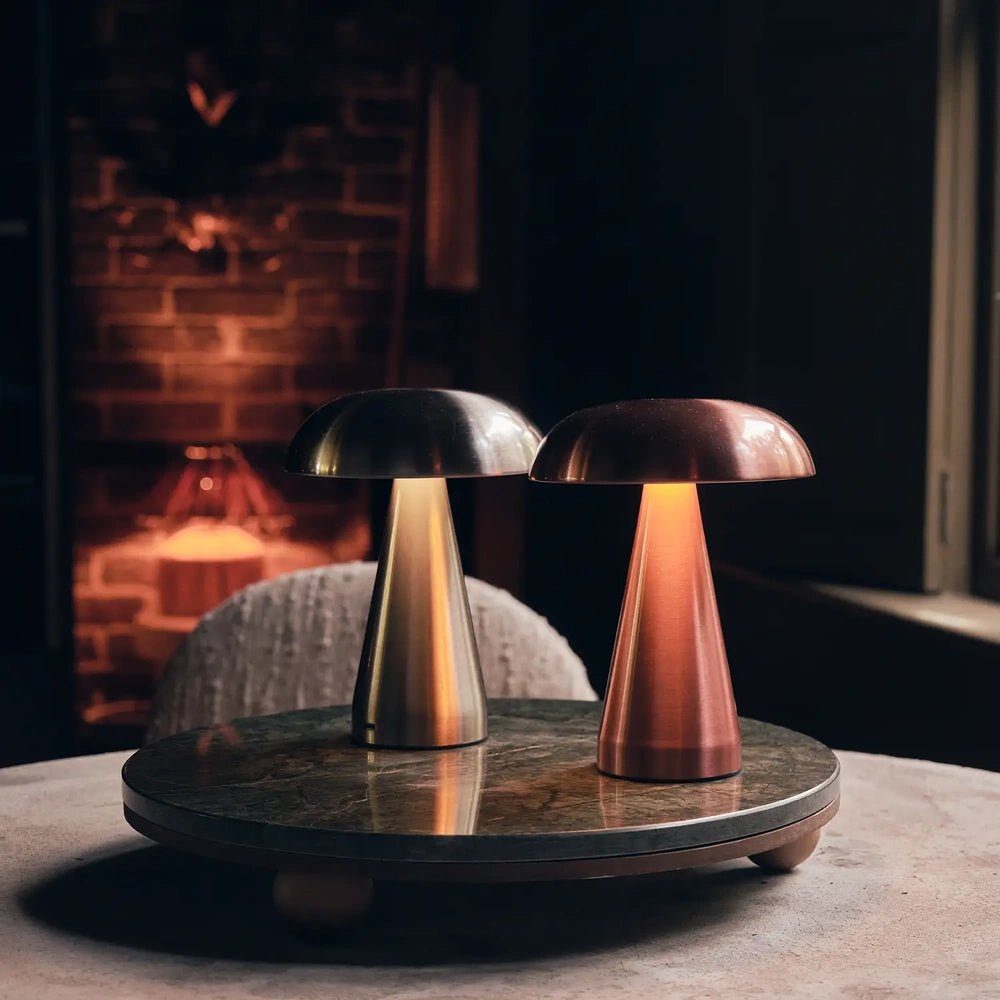 Zenith rose gold portable table lamp shines beside a silver lamp on a round table, showing its reflective domed shade and tapered sculptural base.