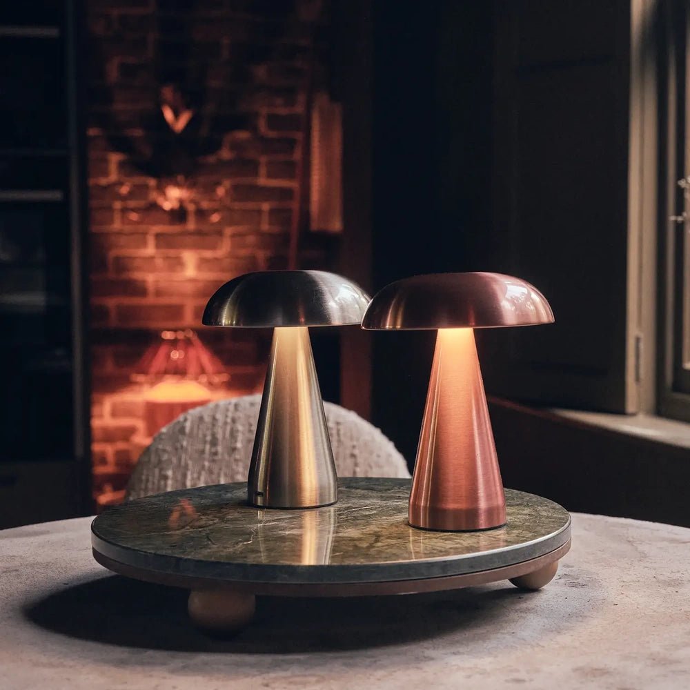 Zenith rose gold portable table lamp with a domed shade and tapered base stands beside a silver lamp on a round table near a fireplace.