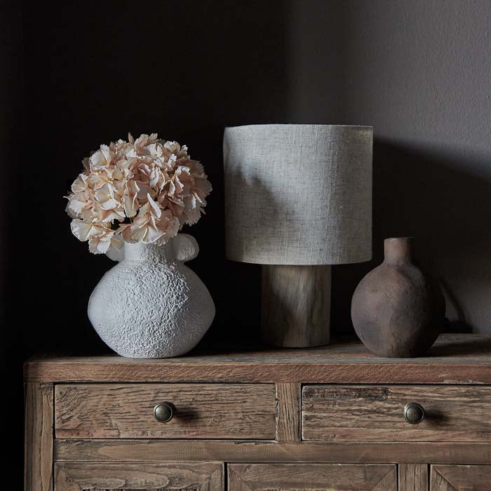 Artificial hydreangea in a round white ecomix vase, displayed on a wooden dresser.