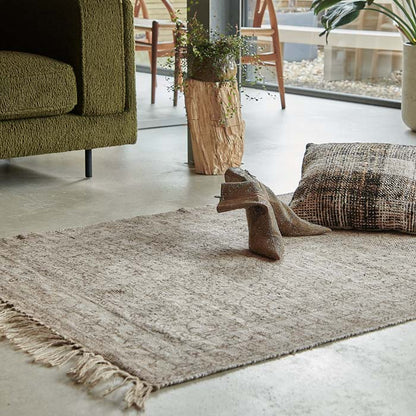 A pale brown woven rug with subtle ornate pattern design and tasseled edges.