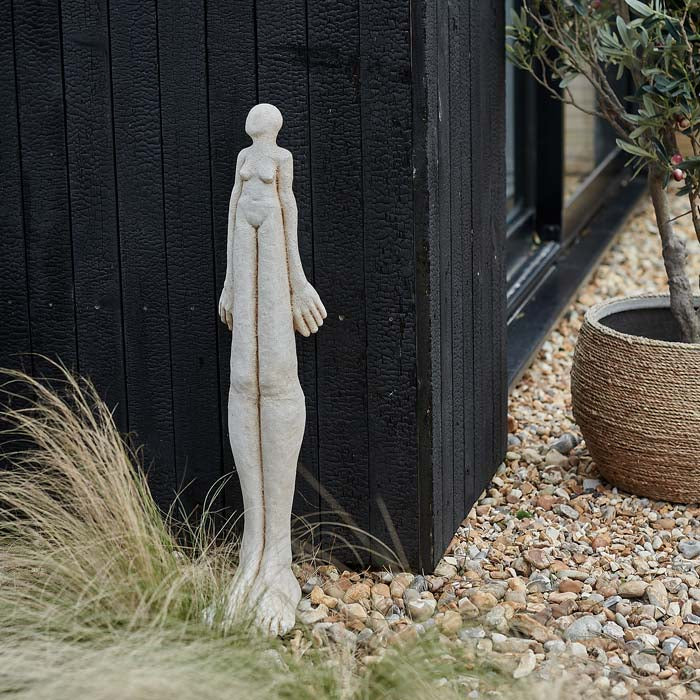 Extra large figurative cement sculpture, with elongated legs, leaning against a wooden wall.