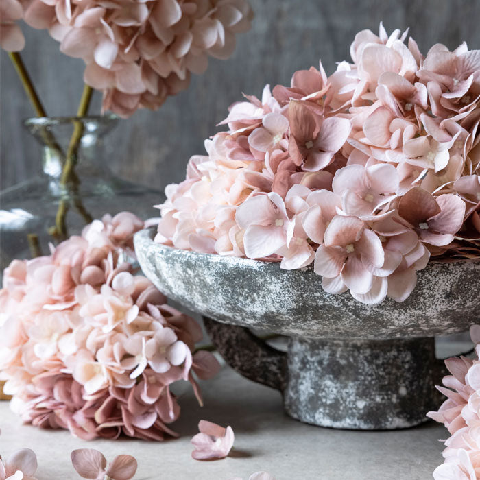 A mound of pink faux hydrangea stems on a large grey bowl surrounded by matching hydrangea stems
