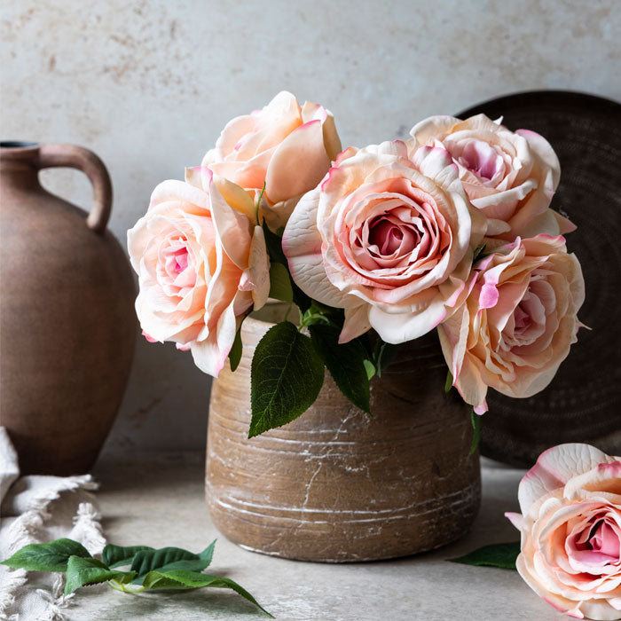 Five artificial blush coloured roses sat in a short brown vase