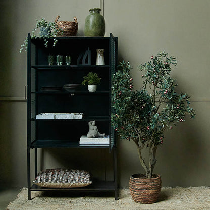 Large artificial olive tree displayed in woven basket next to black iron cabinet.