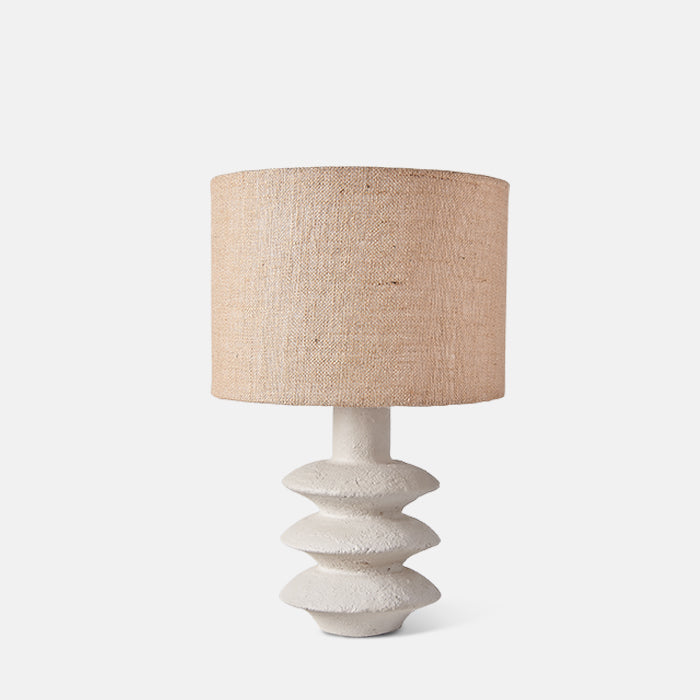 Sculptural white ecomix table lamp with tiered design and drum shade covered in jute fabric.