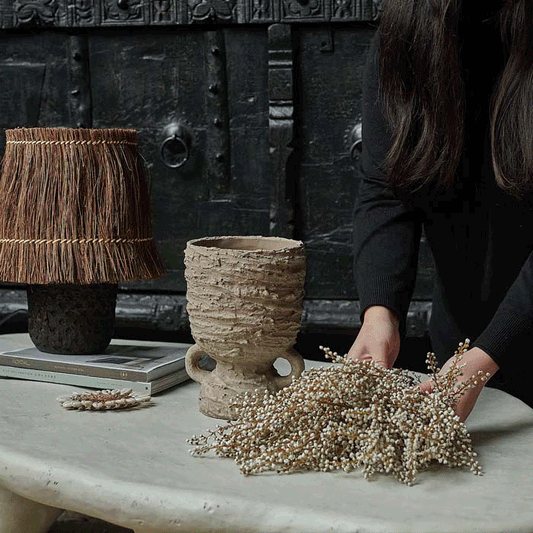 A dark-haired person arranging artificial white heather foliage in a rustic stoneware vase.