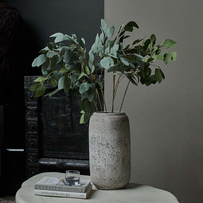 Seven stems of artificial green eucalyptus in a tall grey vase sat on a white table