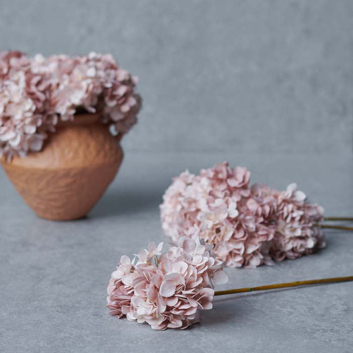 Light pink artificial hydrangea flower stems in front of a coral coloured vase.