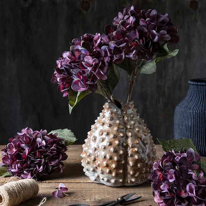 A bobble ceramic vase holding two artificial hydrangea flowers in deep purple tones.