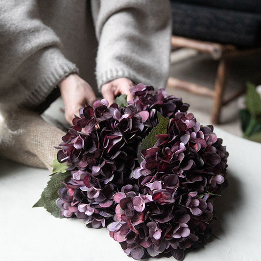 A person's hands tying a bundle of six dark purple artificial hydrangea flowers together.