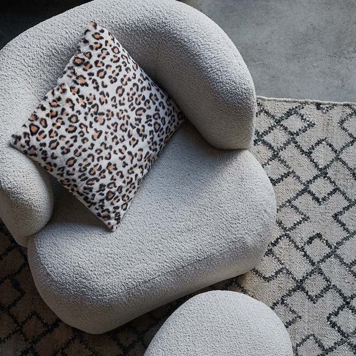 A square white furry cushion with leopard print pattern, on a cream boucle armchair.