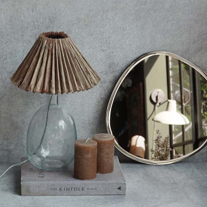 Clear glass lamp with a raffia shade next to two pillar candles and a round mirror