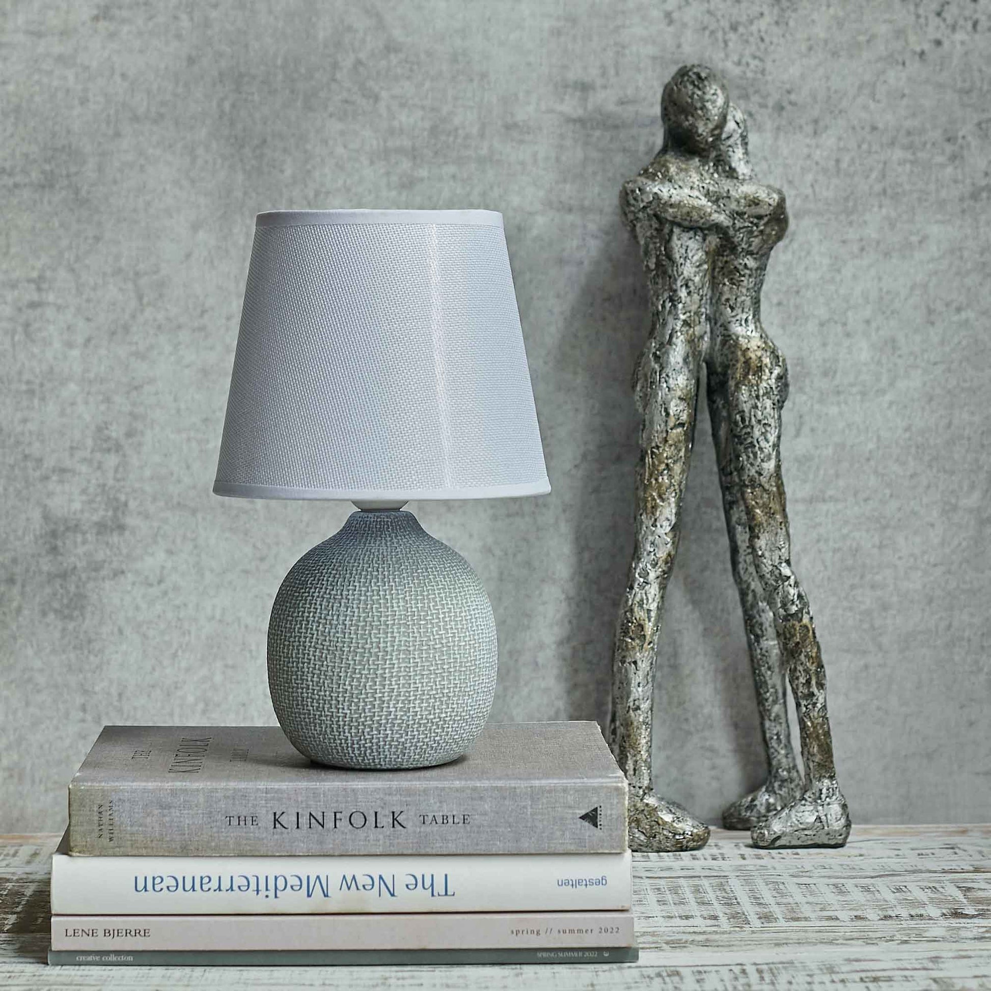 Short round grey table lamp sat on a stack of books in front of two hugging figurines