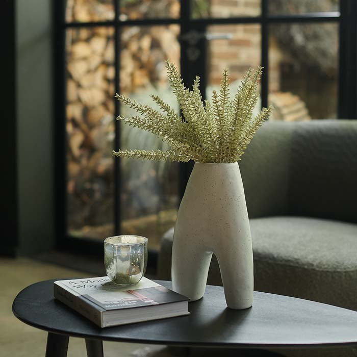 Arch shaped white ceramic vase filled with spiky green stems sat on a black table