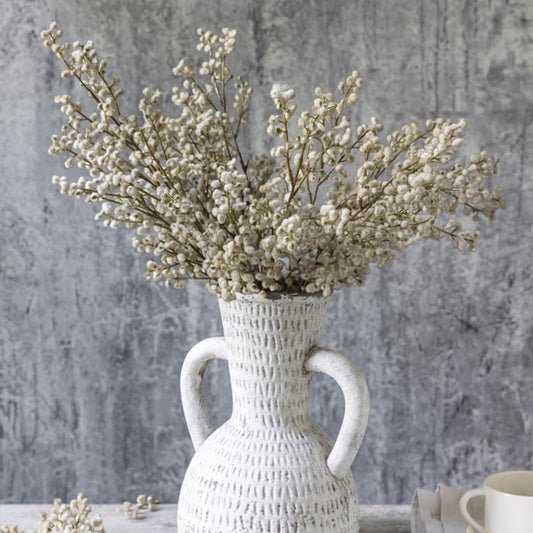 Artificial blush mimosa stems in a distressed-look vase with handles.