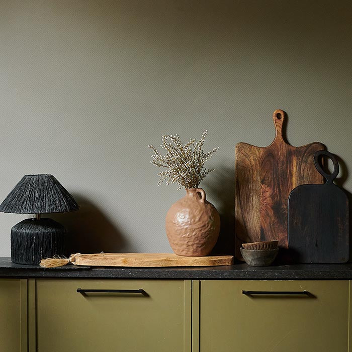 Kitchen worktop filled with three chopping boards, a brown vase and black lamp against a warm taupe painted wall