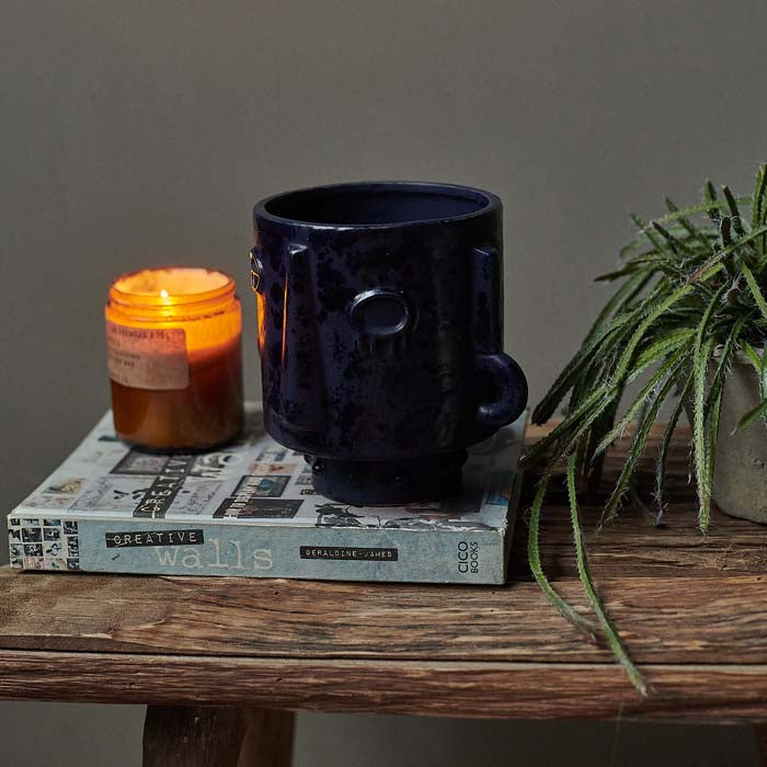 Dark blue plant pot with an abstract face sat on a book next to a lit candle