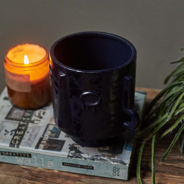 Dark blue round plant pot with a textured abstract face sat on a book next to a candle