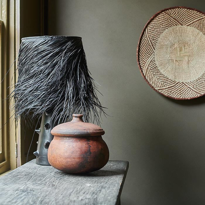 Black lamp and rustic orange pot sat on a wooden table with a warm olive painted wall