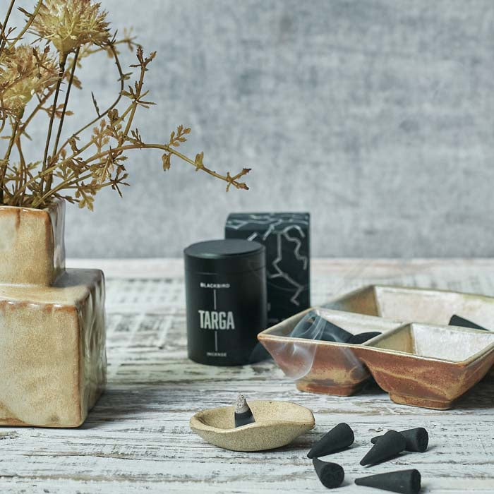 Burning black incense cone sat in a small stoneware dish next to a brown cube vase and tray