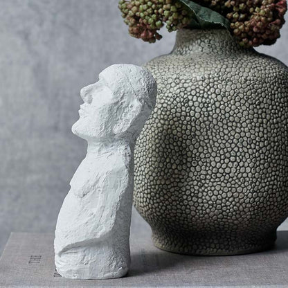 Small white figurine facing to the side next to a textured grey vase
