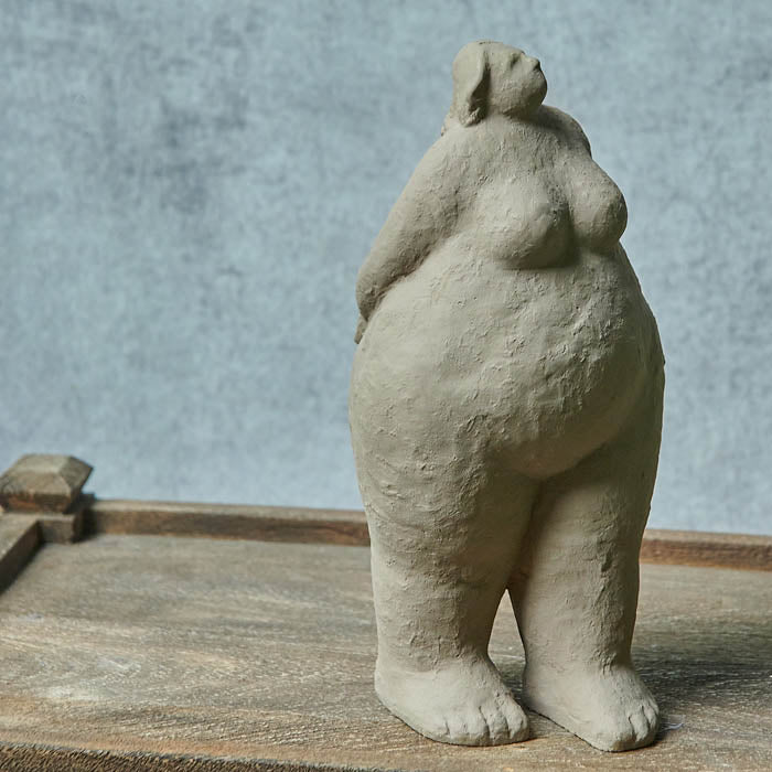 Tall standing female grey cement sculpture looking up with her arms behind her back